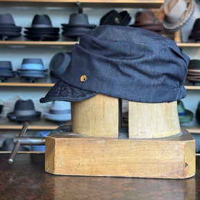 Denim Military Cap in dark blue denim fabric displayed on a wooden hat block with multiple stylish hats in the background, showcasing a vintage-inspired accessory.