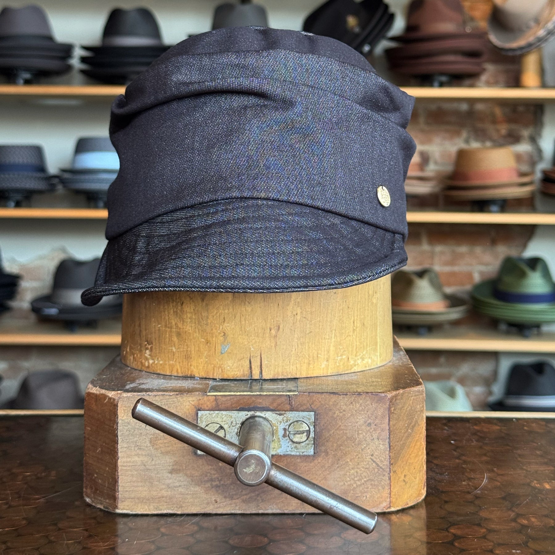 Dark denim fabric Denim Military Cap displayed on a vintage wooden hat block with various hats blurred in the background, highlighting classic style and casual accessory appeal.