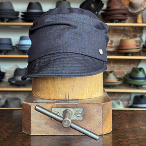 Dark denim fabric Denim Military Cap displayed on a vintage wooden hat block with various hats blurred in the background, highlighting classic style and casual accessory appeal.