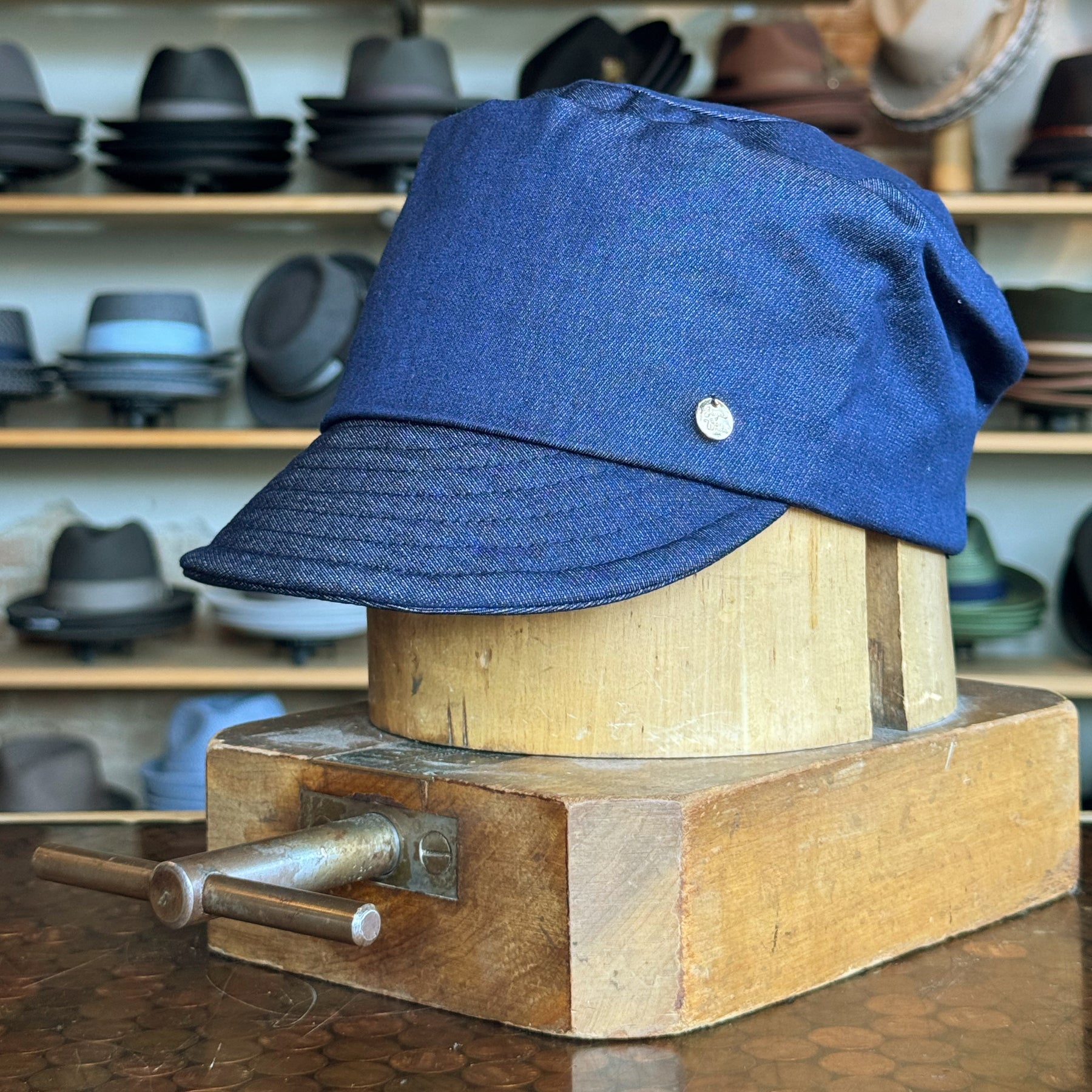 Denim Military Cap in blue displayed on a wooden hat block with shelves of various hats in the background, showcasing a casual and sturdy accessory style.