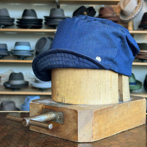Denim Military Cap in blue displayed on a wooden hat block with various hats blurred in the background, highlighting its casual, durable fabric and structured brim style.