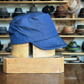 Denim Military Cap displayed on a wooden hat block, featuring a structured brim and deep blue denim fabric, with shelves of various hats blurred in the background.