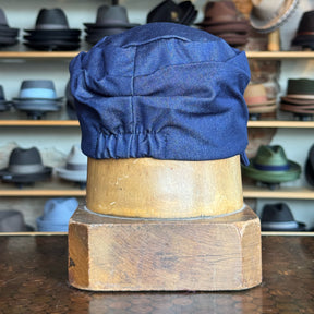 Denim Military Cap displayed on a wooden hat block, showcasing the gathered back detail in blue denim fabric with various hats blurred in the background.