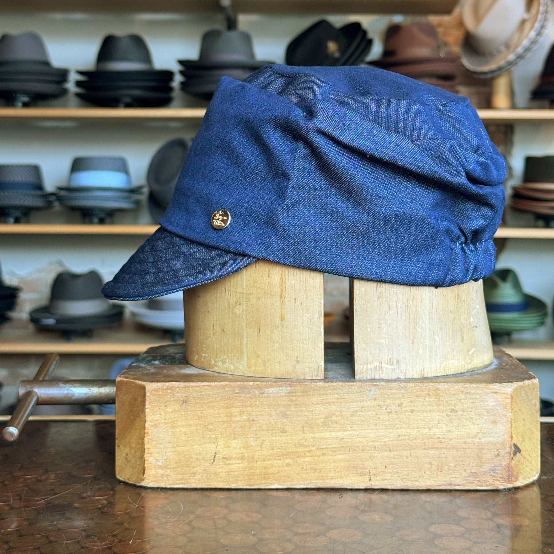 Denim Military Cap in blue denim fabric displayed on a wooden hat block with a small round metal emblem on the side, set against a background of various hats on shelves.