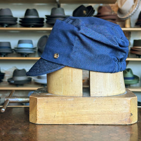 Denim Military Cap in blue denim fabric displayed on a wooden hat block with a small round metal emblem on the side, set against a background of various hats on shelves.
