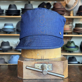 Blue Denim Military Cap displayed on a wooden hat block with various hats blurred in the background, showcasing a classic casual accessory style.