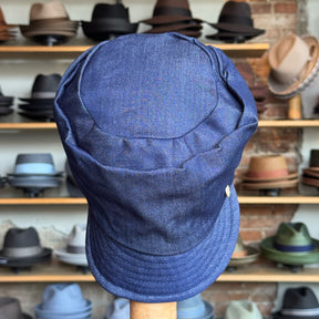 Front view of a blue Denim Military Cap displayed in a hat shop with shelves of various hats in the background.