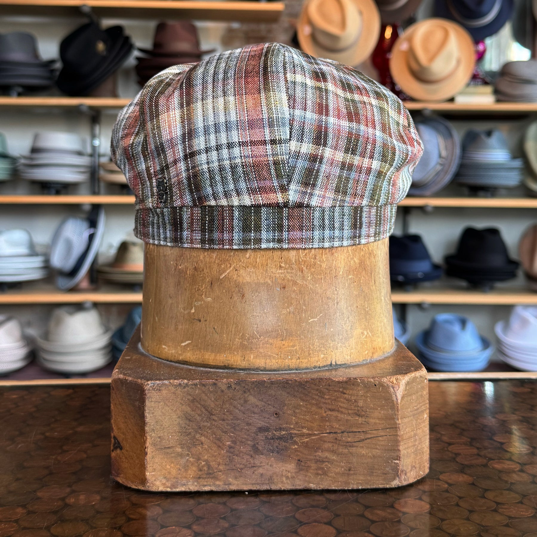Earthling Plaid Hatteras newsboy cap in a multicolor plaid pattern displayed on a wooden hat block with various hats blurred in the background.