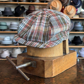 Earthling Plaid Hatteras newsboy cap with a multi-color plaid pattern displayed on a wooden hat block in a shop with various hats in the background.