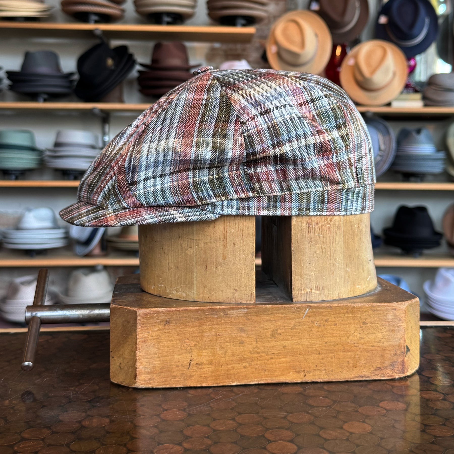 Earthling Plaid Hatteras newsboy cap in multicolor plaid fabric displayed on a wooden hat block with various hats in the background, showcasing classic style and craftsmanship.