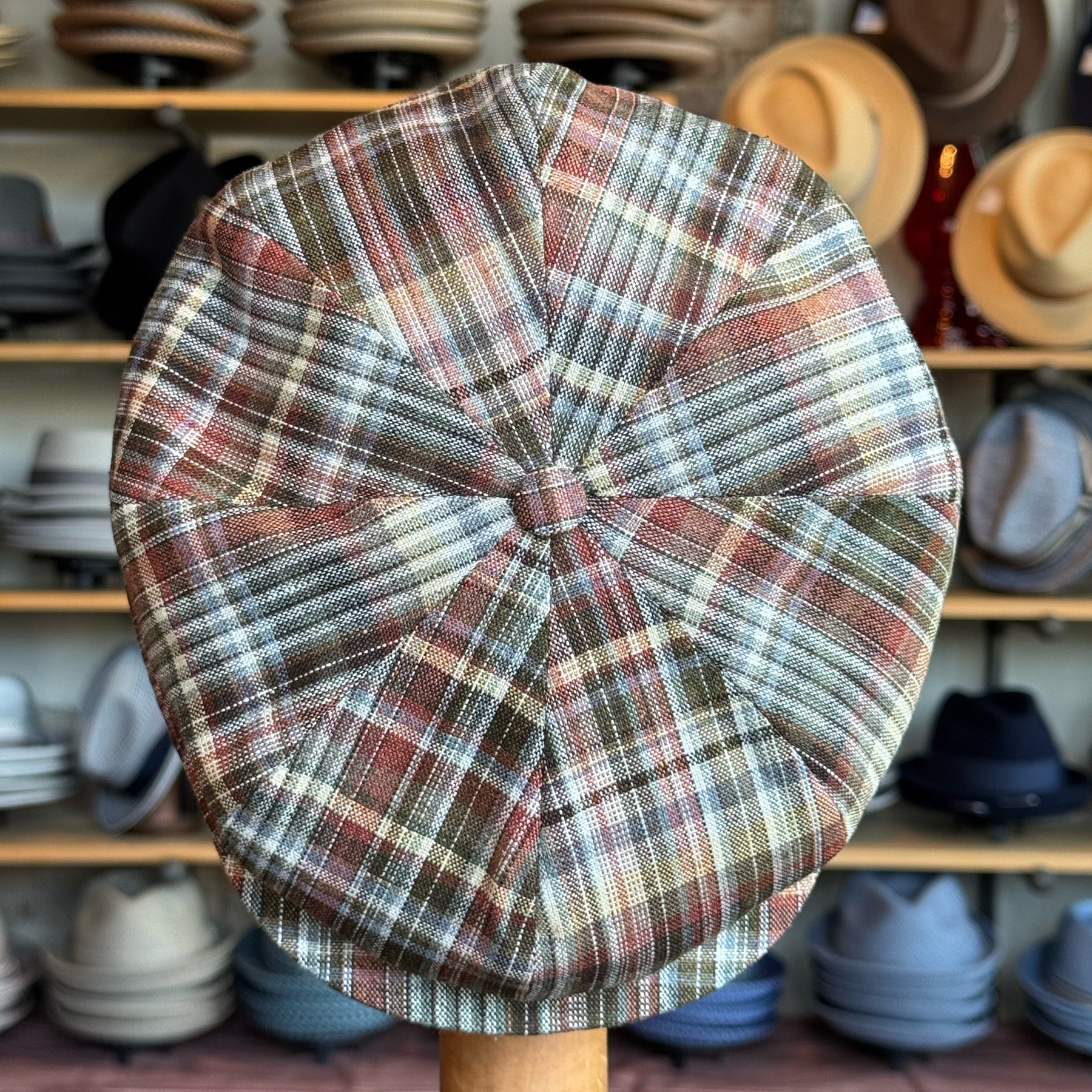 Close-up of Earthling Plaid Hatteras newsboy cap featuring a multicolored plaid pattern in earth tones, displayed in a hat shop with various hats in the background.