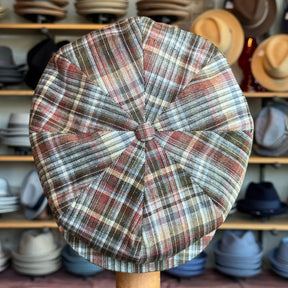 Close-up of Earthling Plaid Hatteras newsboy cap featuring a multicolored plaid pattern in earth tones, displayed in a hat shop with various hats in the background.