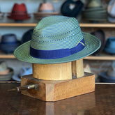 Eli fedora hat in green and blue woven material with a navy blue band, displayed on a wooden hat block in a store with various hats in the background.