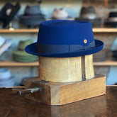 Gareth Pork Pie hat in deep blue wool felt with a black ribbon band and a small round gold emblem, displayed on a wooden hat block with various hats in the background.