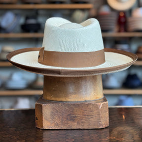 Gaudi Wide Brim Panama Fedora in light woven material with a wide brown band and trim, displayed on a wooden hat stand in a blurred store setting.