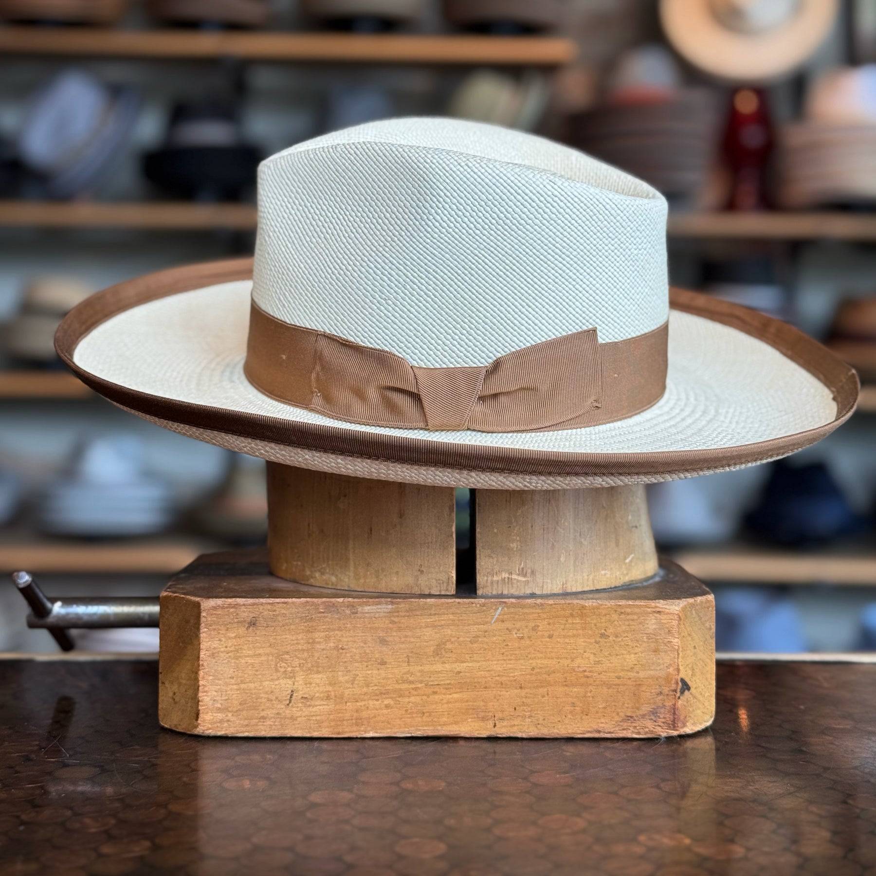Gaudi Wide Brim Panama Fedora with a light woven crown and a tan ribbon band, displayed on a wooden hat block in a vintage hat shop setting.