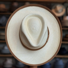 Gaudi Wide Brim Panama Fedora featuring a light woven crown with a wide natural brim trimmed in brown, displayed against a background of various hats.
