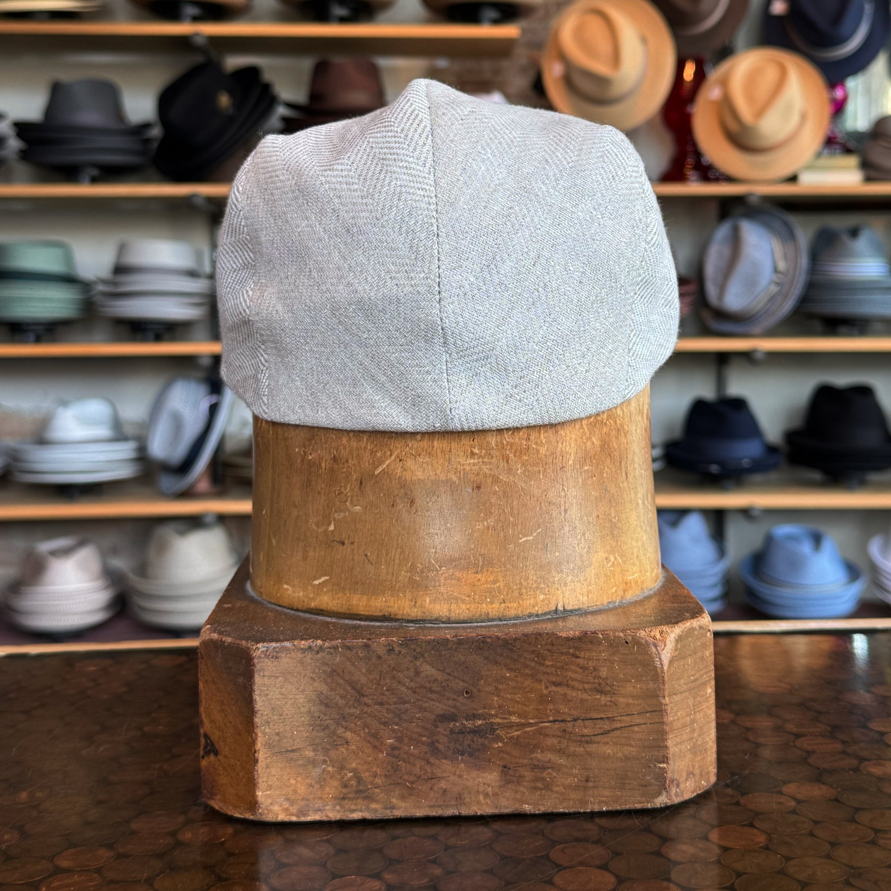 Ghost Herringbone Vintage Flat Cap displayed on a wooden hat block with a subtle herringbone pattern in light gray, surrounded by various hats in the background.