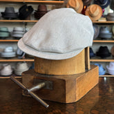 Ghost Herringbone Vintage Flat Cap in light gray with classic herringbone pattern, displayed on a wooden hat block in a shop filled with various hats and caps in the background.