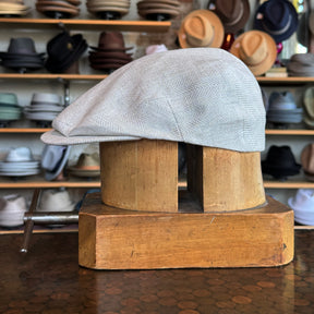 Light gray Ghost Herringbone Vintage Flat Cap displayed on a wooden hat block with shelves of various hats in the background.