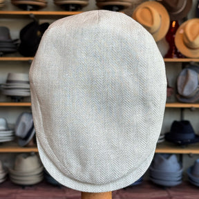 Ghost Herringbone Vintage Flat Cap displayed on a stand, featuring a subtle herringbone pattern in light neutral tones, surrounded by various hats in the background.