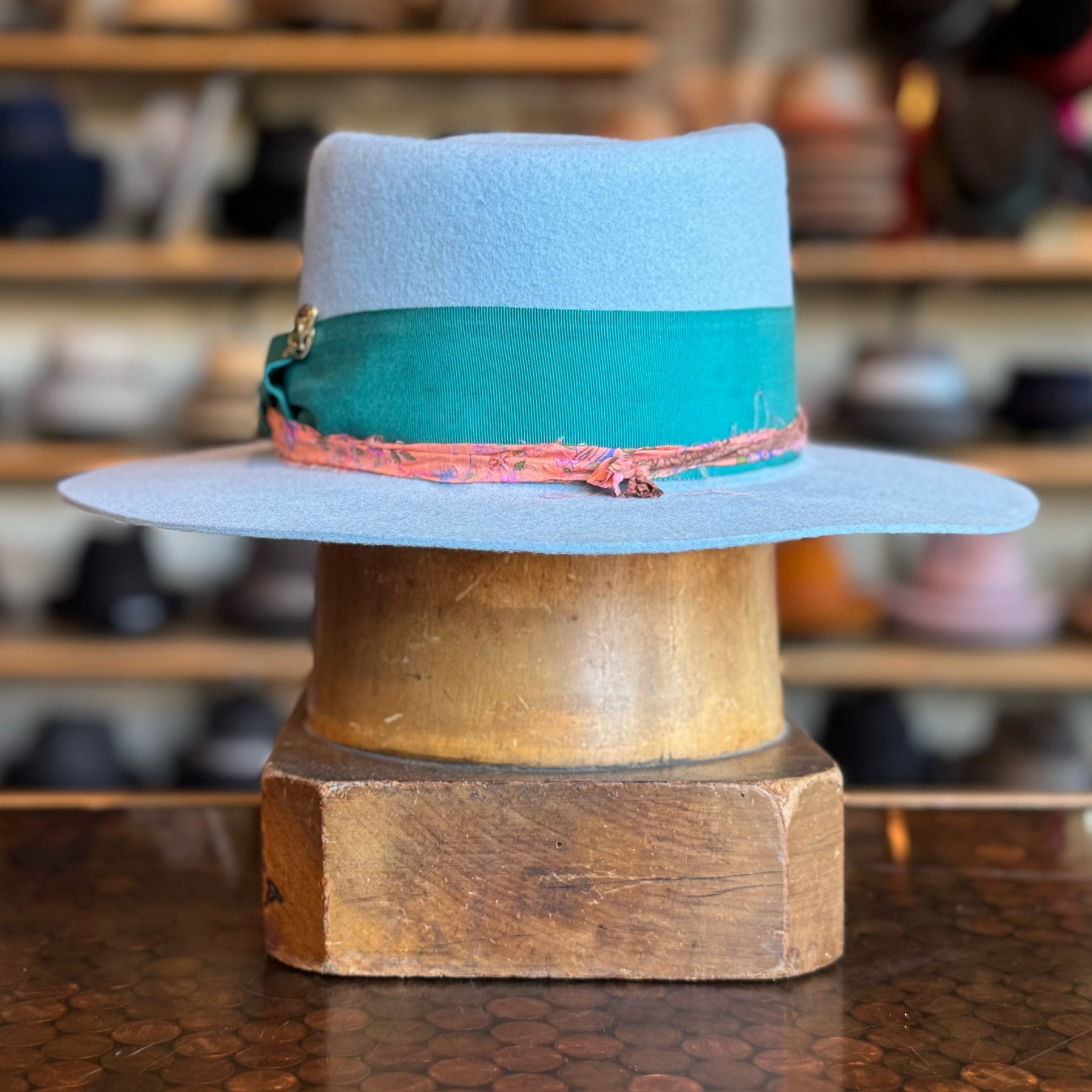 Gin Tansy Flat Brim Fedora in soft baby blue with a wide teal band and a textured coral accent, displayed on a wooden hat block with blurred shelves of hats in the background.