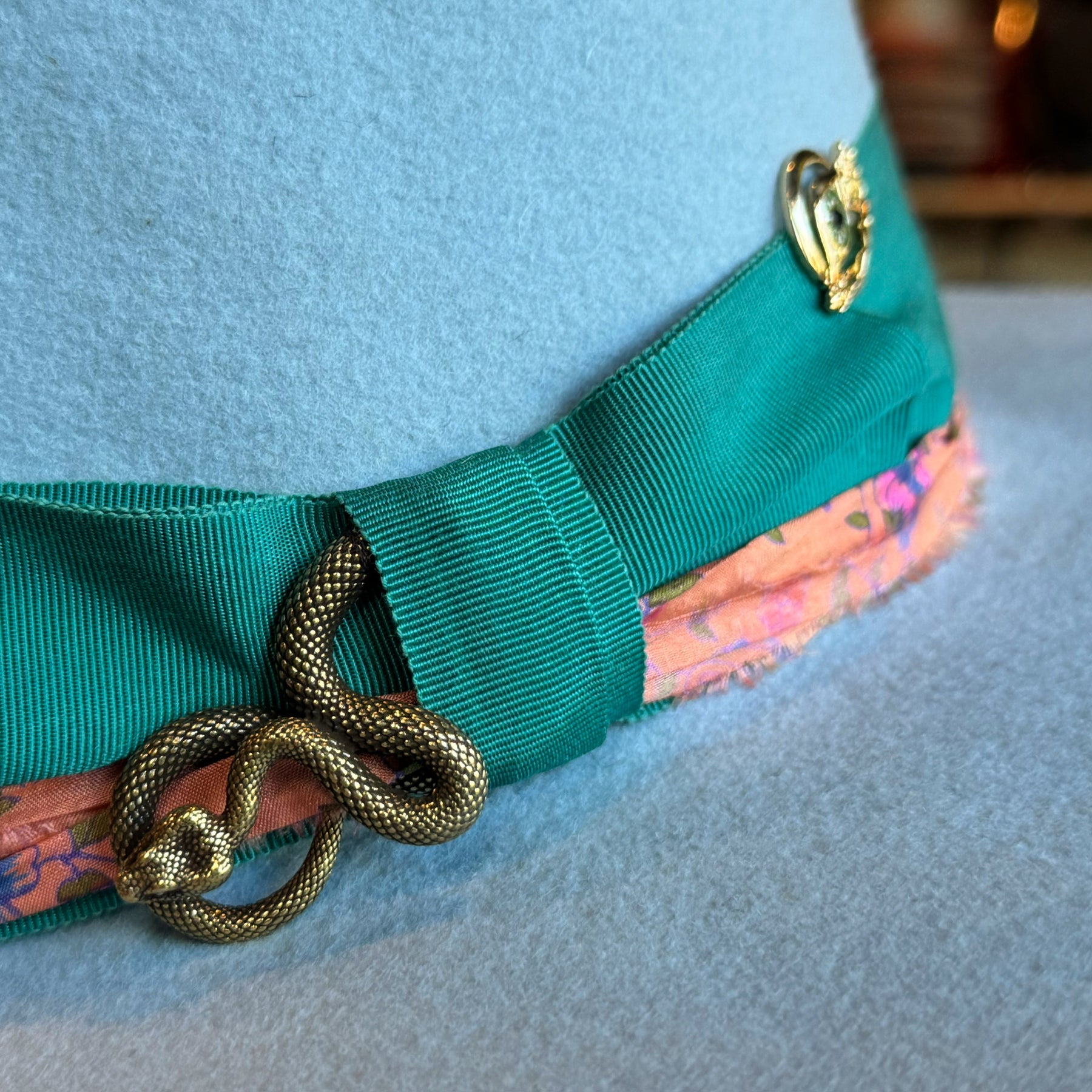 Close-up of a baby blue Gin Tansy Flat Brim Fedora featuring a green grosgrain ribbon band, a decorative bronze snake pin, and a floral patterned lining detail.