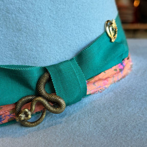 Close-up of a baby blue Gin Tansy Flat Brim Fedora featuring a green grosgrain ribbon band, a decorative bronze snake pin, and a floral patterned lining detail.