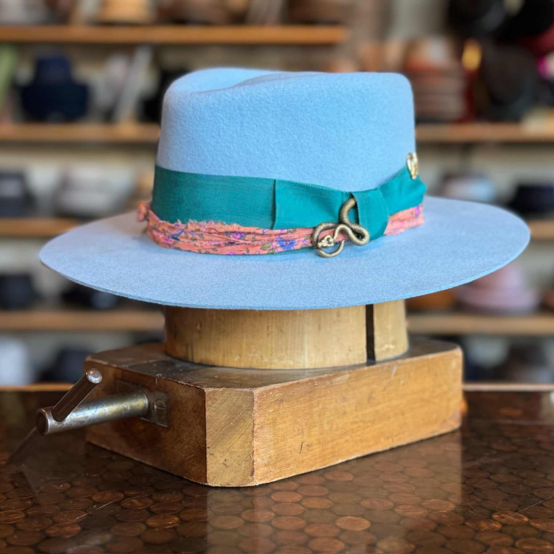 Gin Tansy Flat Brim Fedora in baby blue with a wide teal ribbon and decorative gold accents, displayed on a wooden hat block in a hat shop setting.