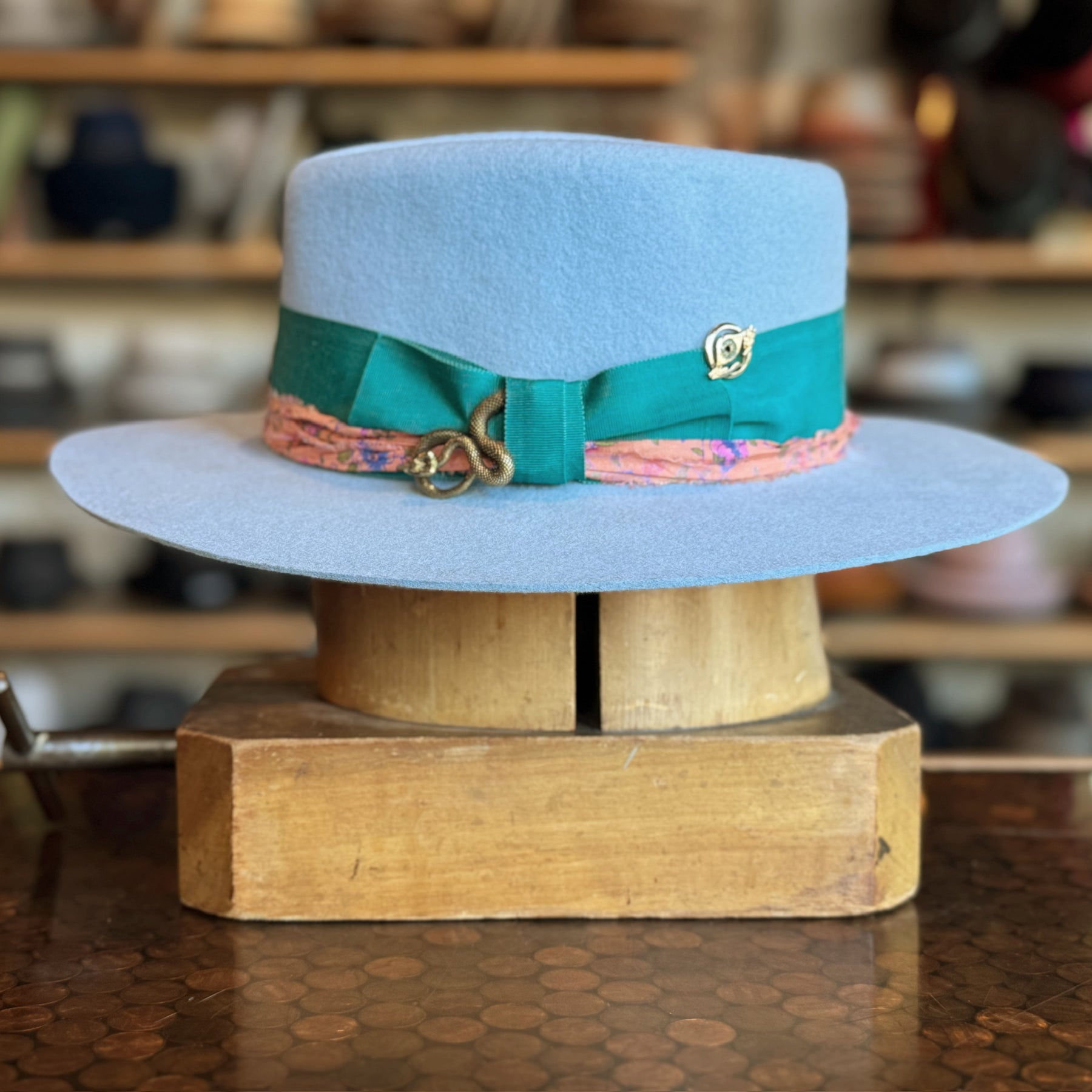 Gin Tansy Flat Brim Fedora in soft baby blue felt with a green ribbon band accented by a pink patterned trim and a decorative gold pin, displayed on a wooden stand.