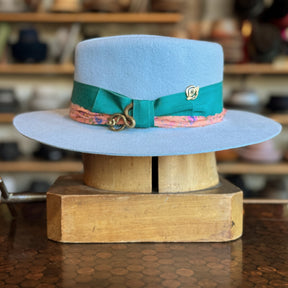 Gin Tansy Flat Brim Fedora in soft baby blue felt with a green ribbon band accented by a pink patterned trim and a decorative gold pin, displayed on a wooden stand.