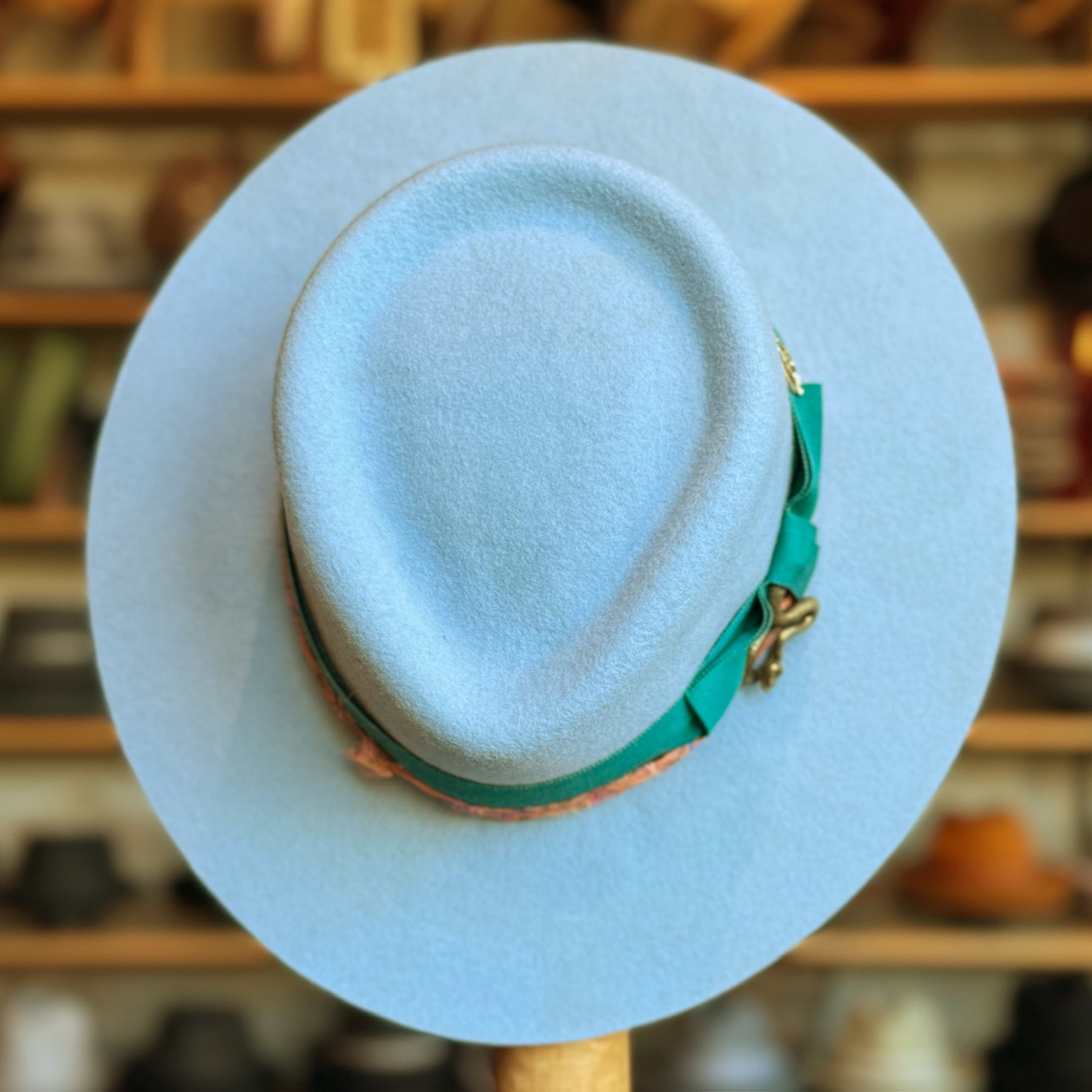 Light blue Gin Tansy Flat Brim Fedora with a green ribbon band and gold accent, displayed on a stand with a blurred background of hats on shelves.
