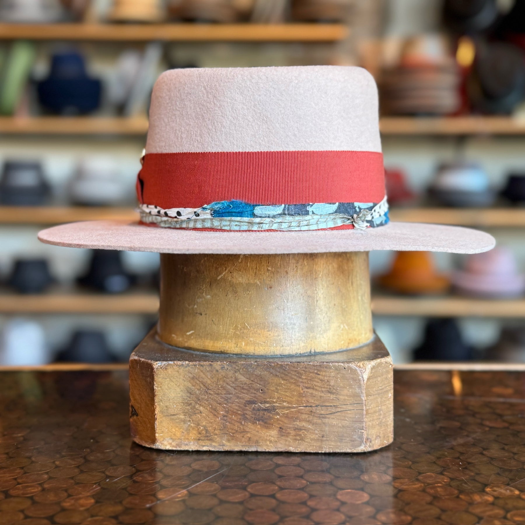 Gin Tansy Flat Brim Fedora in light pink with a wide red band and a patterned trim, displayed on a wooden hat block with shelves of various hats blurred in the background.