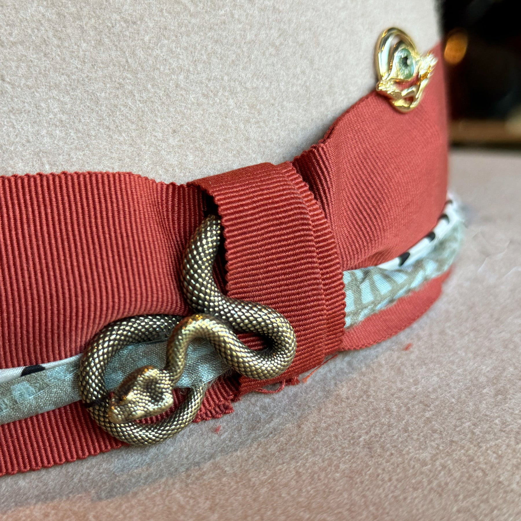 Close-up detail of a beige Gin Tansy Flat Brim Fedora featuring a textured red ribbon band, an intertwined metallic snake accessory, and a patterned fabric accent beneath the band.