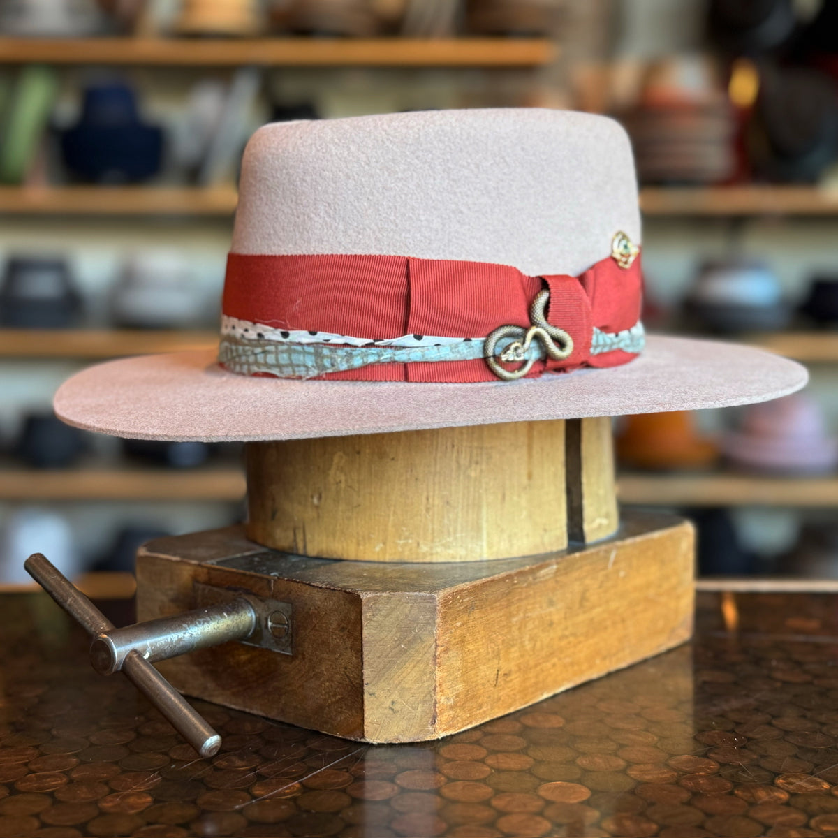 Gin Tansy Flat Brim Fedora in soft light pink with a wide orange band and decorative silver accents, displayed on a wooden hat block in a hat shop setting.