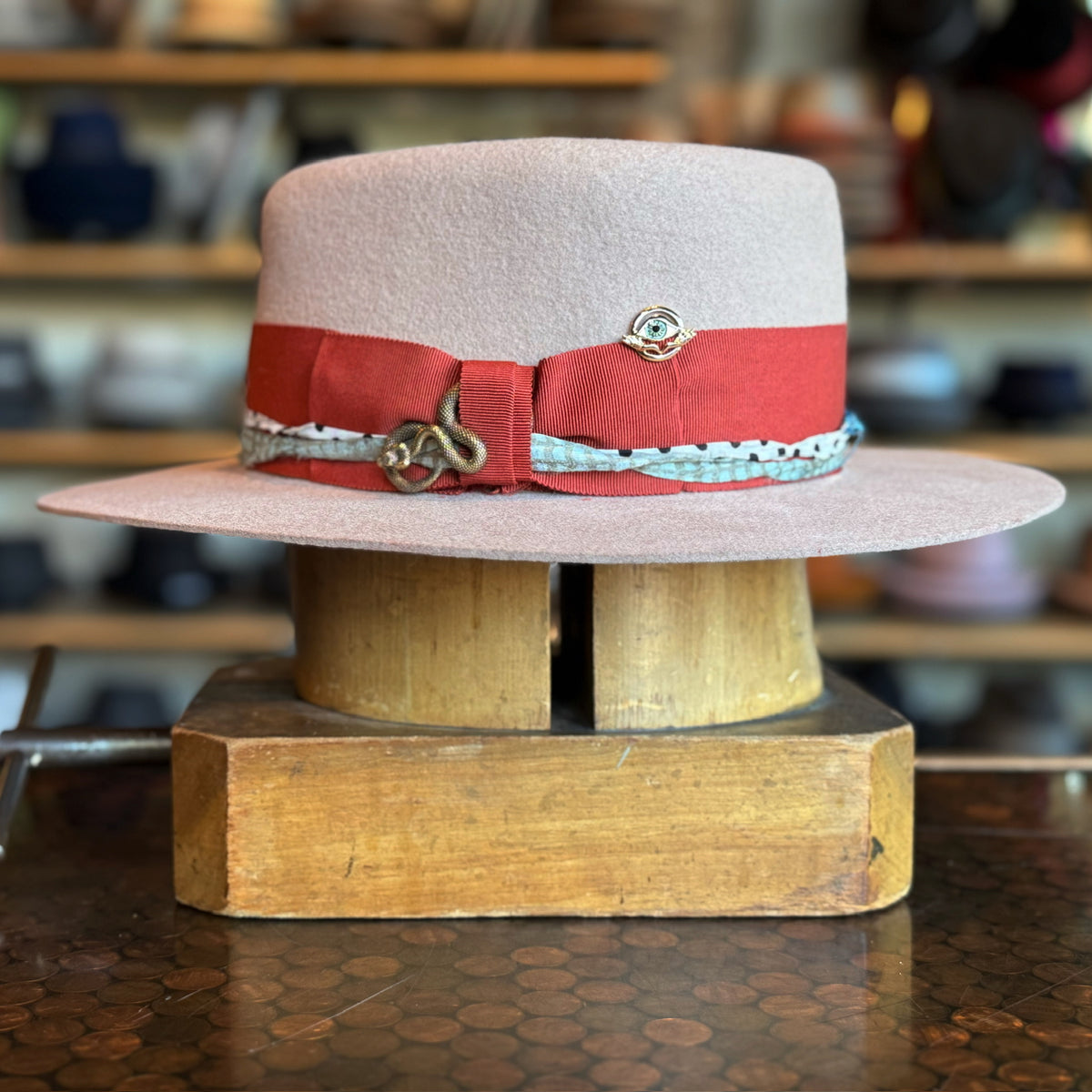 Gin Tansy Flat Brim Fedora in a soft pale pink shade with a wide flat brim, featuring a bold red band accented with a decorative silver charm and a patterned trim.