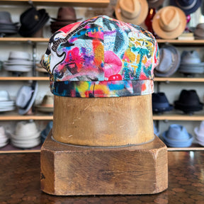 Colorful graffiti-style design on a fabric newsboy cap displayed on a wooden hat block in a hat shop setting, showcasing the Graffiti Hatteras flat cap.
