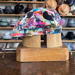 Graffiti Hatteras cap in vibrant multicolor graffiti print with a flat cap style, displayed on a wooden hat block with various hats blurred in the background.