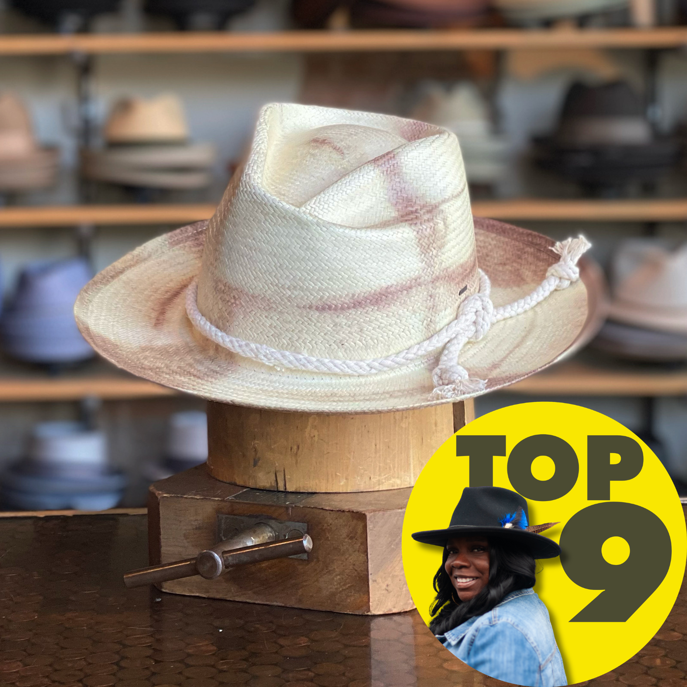 Hinx white and beige straw fedora with twisted rope band displayed on a wooden stand, surrounded by shelves of various hats in the background.