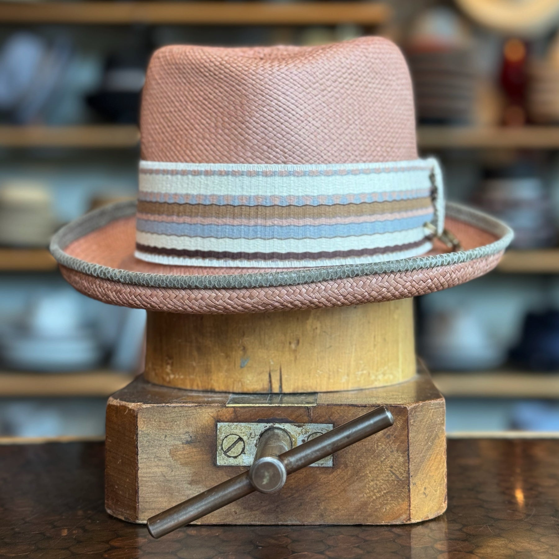 Honey Truffle Legba flat cap with a textured woven design in soft pink tones and a striped fabric band in muted blue and cream, displayed on a wooden hat block.
