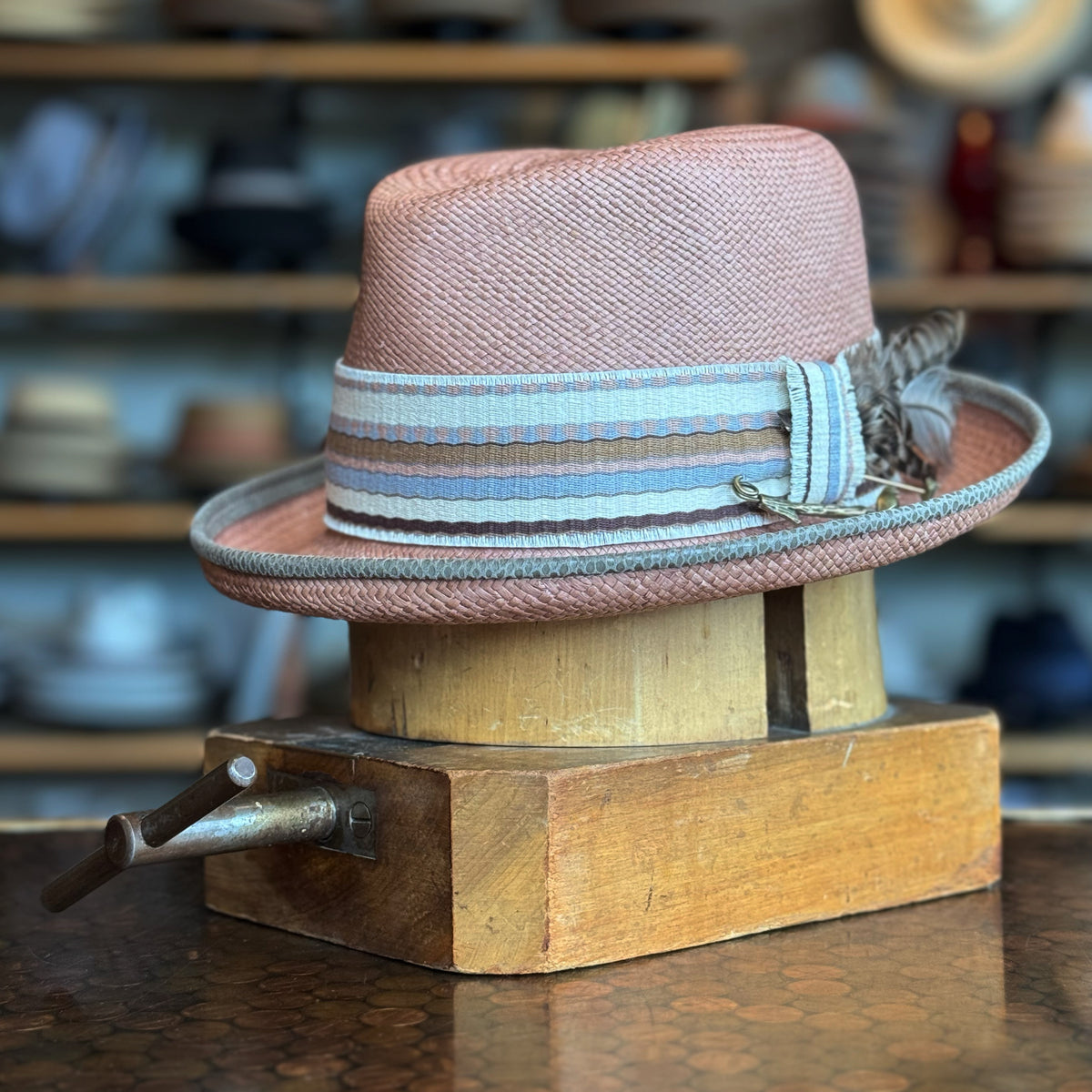 Honey Truffle Legba hat in soft pink with a wide striped blue and beige ribbon and decorative feather, displayed on a wooden hat block in a vintage shop setting.