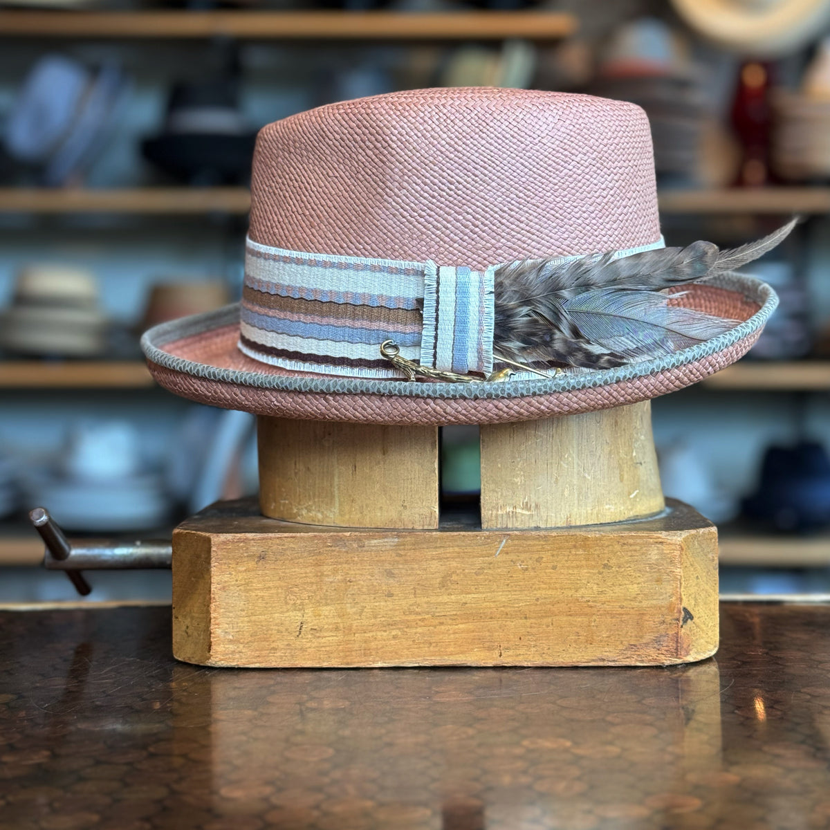 Honey Truffle Legba flat cap in a soft pink woven material with a striped ribbon band and decorative feathers, displayed on a wooden hat block against a blurred background of hats.
