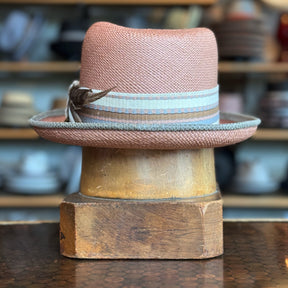 Honey Truffle Legba straw fedora hat with a light beige and blue striped band and decorative feather, displayed on a wooden hat block in a vintage shop setting.