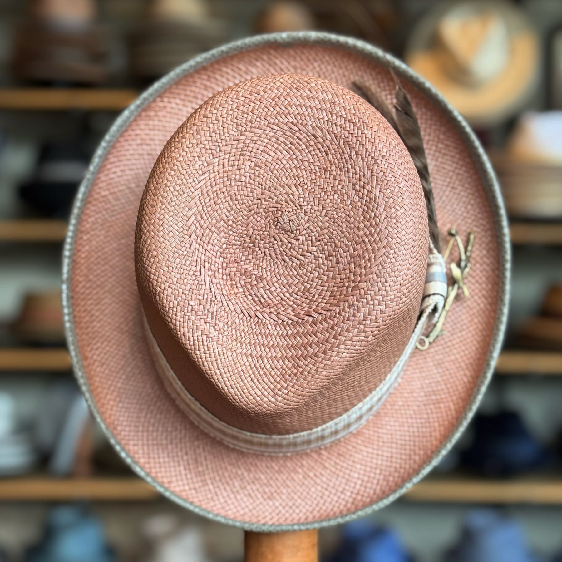 Honey Truffle Legba natural woven straw hat with a light beige band and decorative feather, displayed on a wooden stand against a blurred background of various hats.