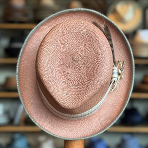 Honey Truffle Legba natural woven straw hat with a light beige band and decorative feather, displayed on a wooden stand against a blurred background of various hats.