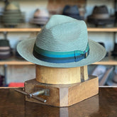 Side-Eye fedora hat made from woven natural fibers with a green and blue striped hatband, displayed on a wooden hat block with shelves of various hats in the background.