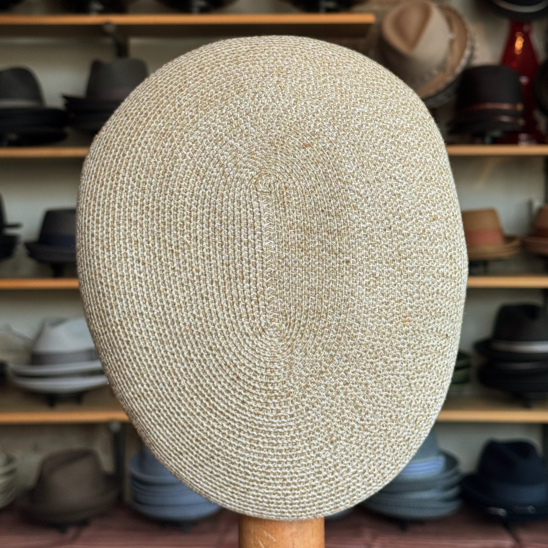 Close-up of a beige woven Dan Ascot Cap displayed on a wooden stand, showcasing its detailed texture with various hats blurred in the background.