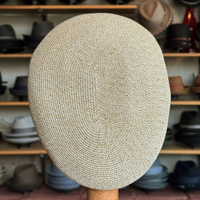 Close-up of a beige woven Dan Ascot Cap displayed on a wooden stand, showcasing its detailed texture with various hats blurred in the background.