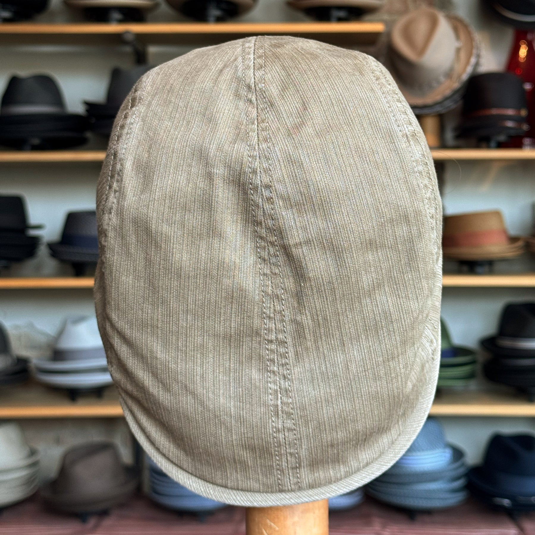 Beige corduroy flat cap displayed on a wooden stand with a background of various hats on shelves, showcasing the Leland Two-Panel Ivy Cap style.