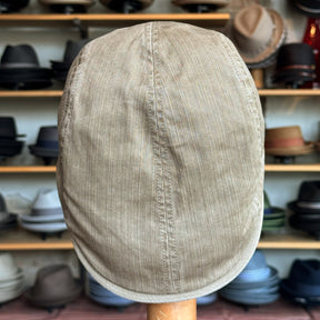 Beige corduroy flat cap displayed on a wooden stand with a background of various hats on shelves, showcasing the Leland Two-Panel Ivy Cap style.